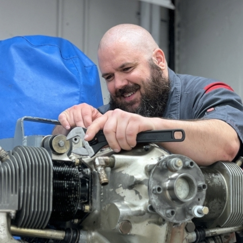 smiling aviation maintenance student uses a wrench to work on an aircraft engine during hands-on training in a lab setting. The student is focused on adjusting engine components while wearing a gray work uniform, highlighting Greenville Technical College’s aviation maintenance program and real-world technical training.