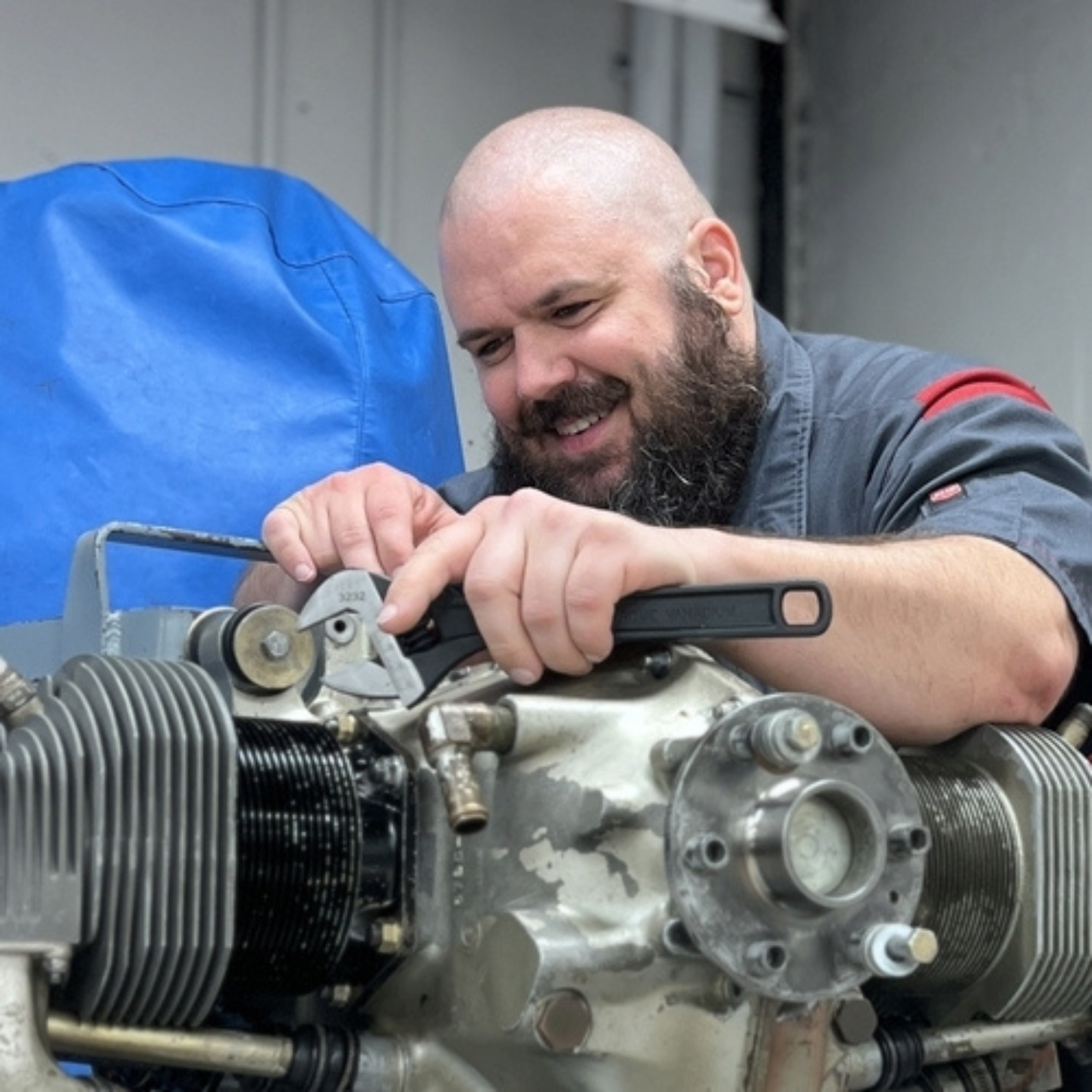 smiling aviation maintenance student uses a wrench to work on an aircraft engine during hands-on training in a lab setting. The student is focused on adjusting engine components while wearing a gray work uniform, highlighting Greenville Technical College’s aviation maintenance program and real-world technical training.
