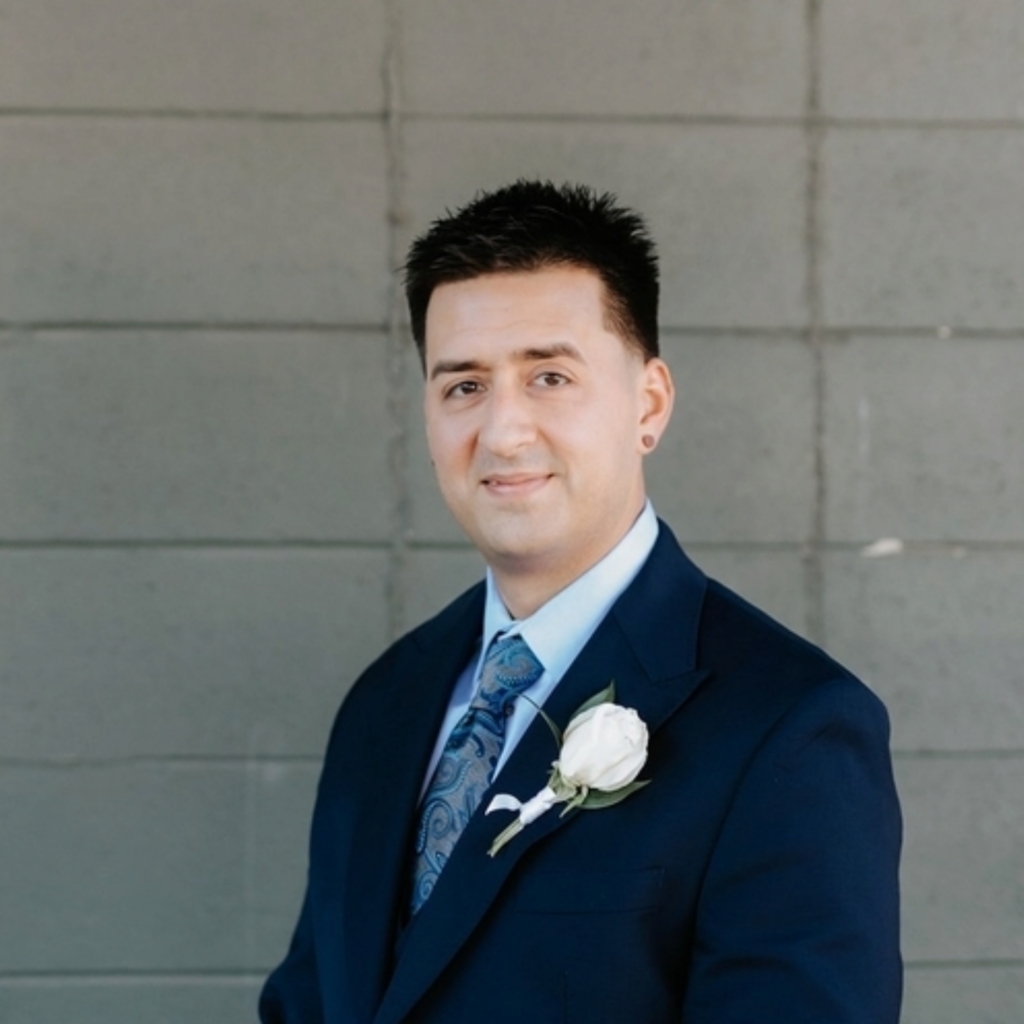 William, Greenville Technical College student wearing a navy blue suit, light blue shirt, and patterned blue tie poses in front of a gray tiled wall. He is wearing a white rose boutonniere and smiling slightly at the camera.
