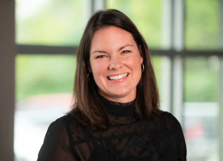 Gina Blohm, Executive Director of the Greenville Tech Foundation, smiling in a professional headshot. She is wearing a black blouse and standing in front of large windows with a softly blurred green outdoor background.