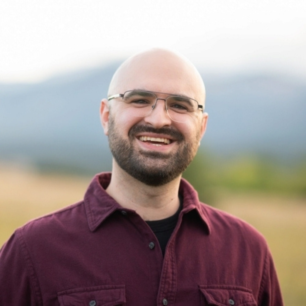 Andrew smiles at the camera while standing outdoors in front of a softly blurred field and mountain landscape. He is wearing glasses, a burgundy button-up shirt over a black T-shirt, and has a neatly trimmed beard. The natural background and warm lighting create a welcoming portrait.