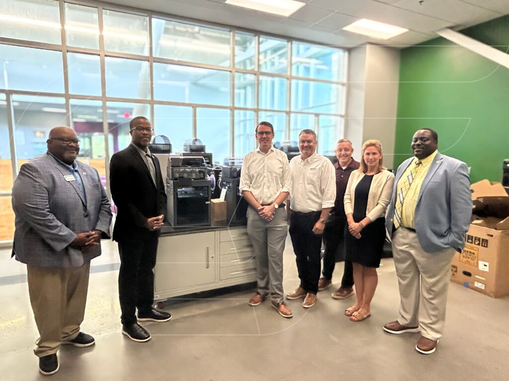 Representatives from Greenville Technical College and the Bosch Community Fund stand together in the Advanced Manufacturing lab beside newly installed manufacturing equipment funded by a Bosch Community Fund grant. The group poses in front of large windows inside the lab, highlighting the partnership supporting hands-on workforce training for students.