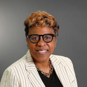 Professional headshot of Babette Jones smiling, wearing black glasses, a cream pinstripe blazer, black top, and gold chain necklace against a gray background.