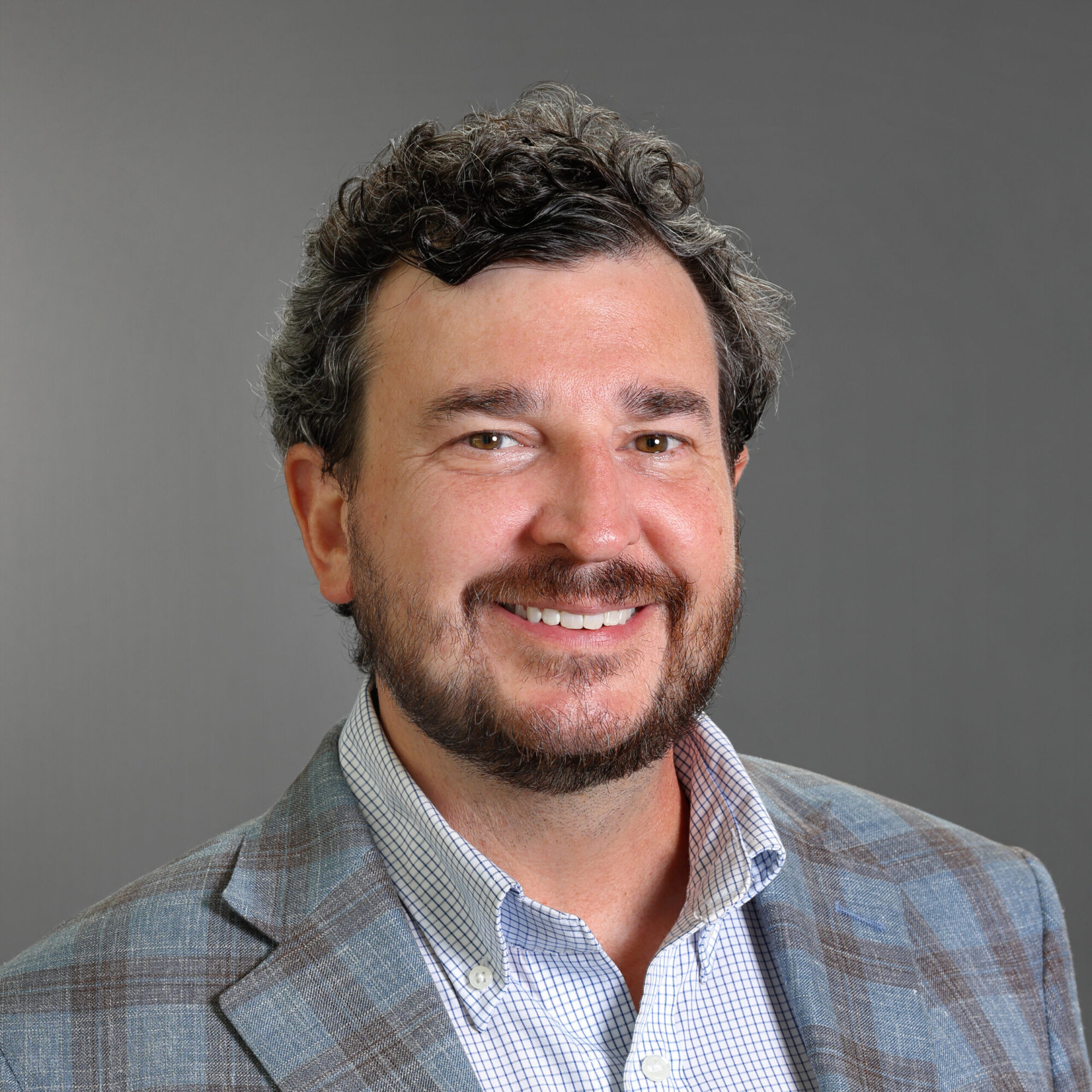 Professional headshot of Brandon Scott smiling, wearing a gray plaid blazer and light checkered dress shirt against a gray background.