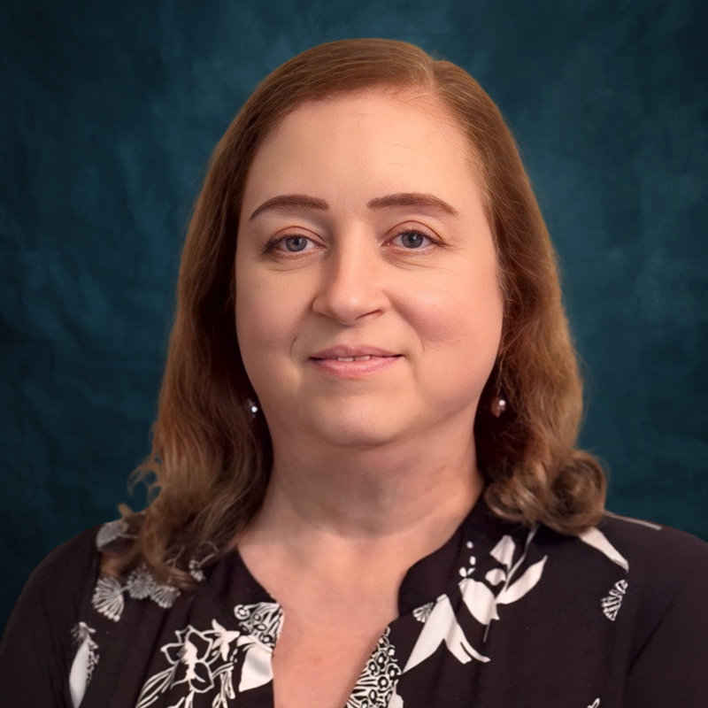 Professional headshot of Cindy Bayles. She is smiling and wearing a black blouse with a white floral pattern against a dark blue background.