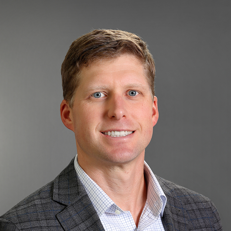 Professional headshot of Drew Boland smiling, wearing a dark gray blazer over a light checkered dress shirt against a gray background.