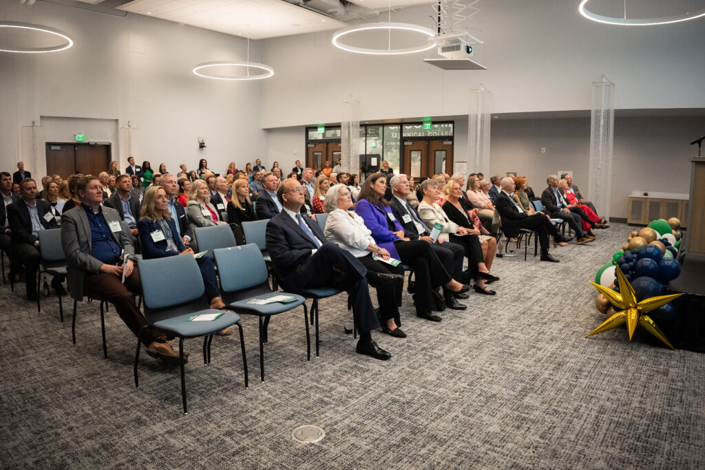 Audience members attend a Greenville Tech Foundation event, seated and listening attentively to a speaker in a modern conference space decorated with blue, green, white, and gold balloon arrangements.