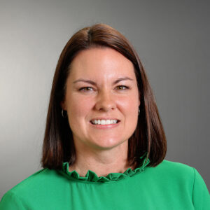 Professional headshot of Gina Blohm smiling, wearing a bright green blouse with a ruffled neckline against a gray background.