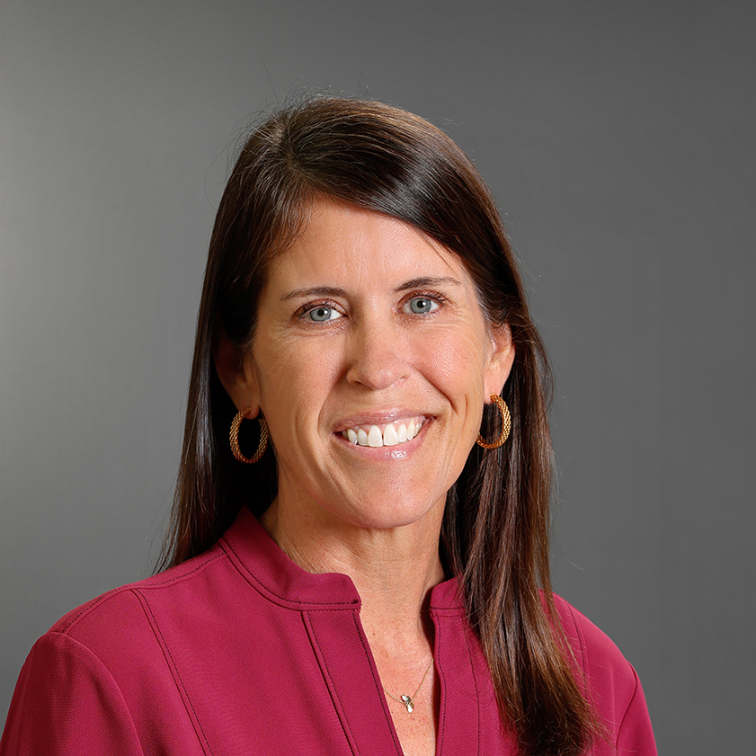 Professional headshot of Katie Howell smiling, wearing a burgundy blouse, gold hoop earrings, and a delicate necklace against a gray background.