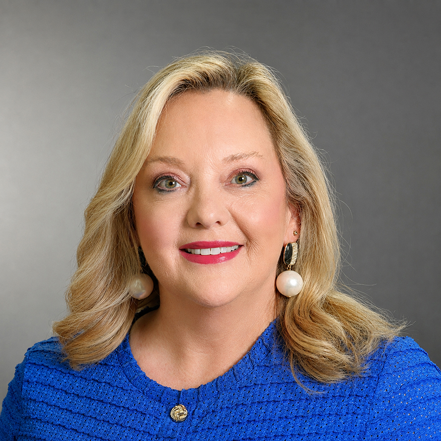 Professional headshot of Kimberly Shell smiling, wearing a bright blue textured top and statement pearl earrings against a gray background.