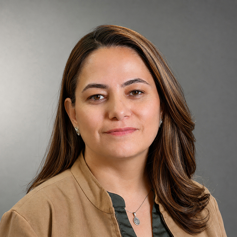 Professional headshot of Maria Duque wearing a tan jacket over a dark top, with shoulder-length highlighted brown hair, against a gray background.