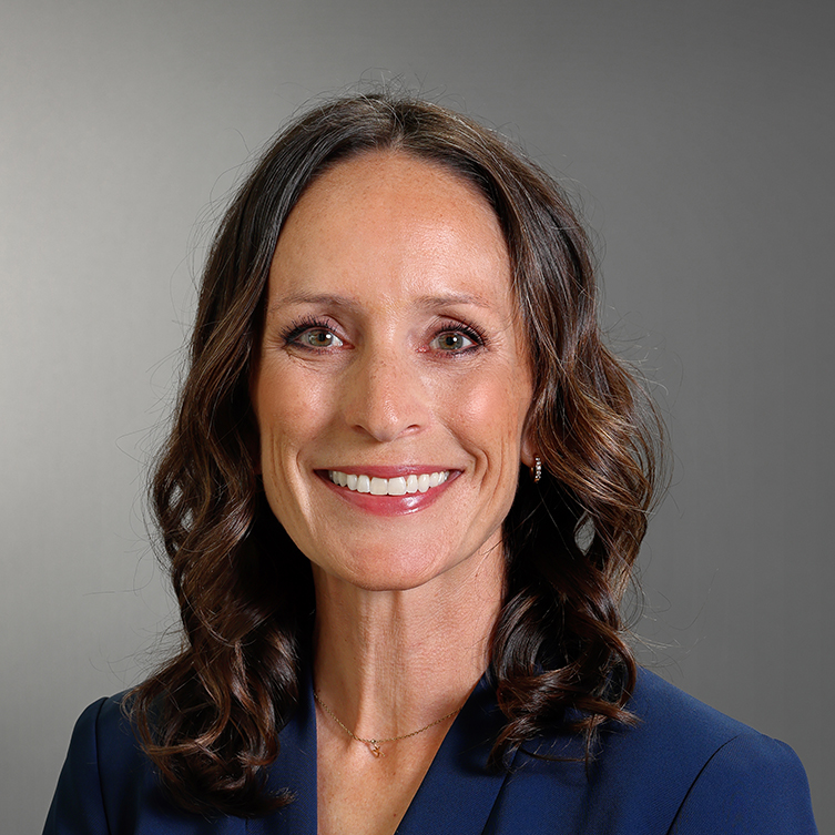 Professional headshot of Megan Finnern smiling, wearing a navy blazer with shoulder-length wavy brown hair against a gray background.