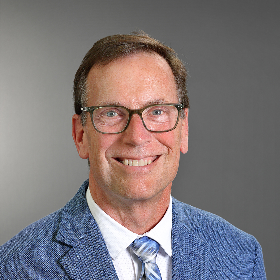 Professional headshot of Mike McCormick smiling, wearing glasses, a blue blazer, white dress shirt, and blue patterned tie against a gray background.