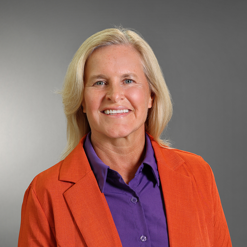 Professional headshot of Stephanie Ferguson smiling, wearing an orange blazer over a purple collared shirt against a gray background.