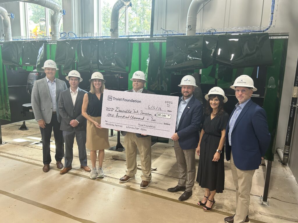Representatives from Truist Foundation and the Greenville Tech Foundation stand together wearing white hard hats in Greenville Technical College's welding facility while holding a ceremonial $100,000 donation check. The gift will support the Center for Welding and Automation Excellence, helping provide students with hands-on training and industry-relevant learning opportunities.
