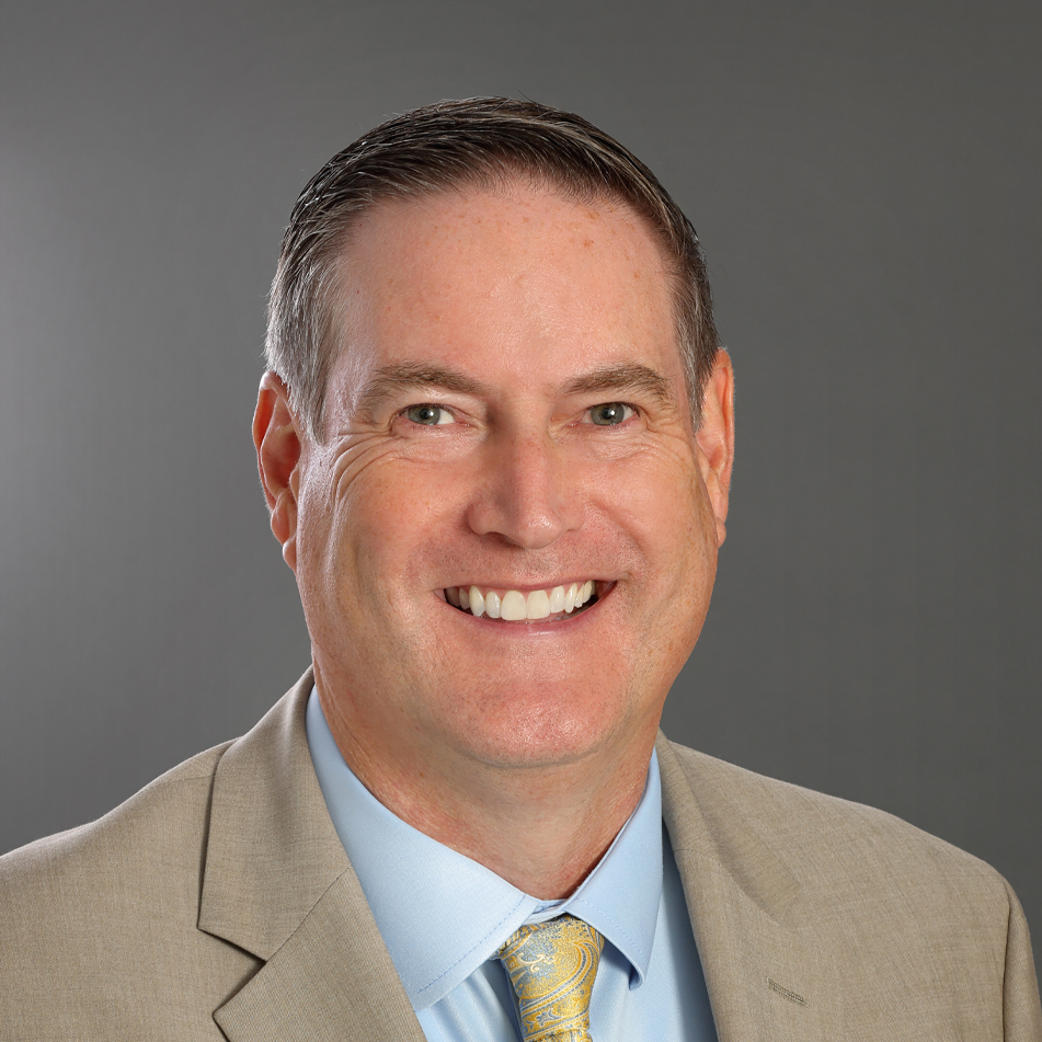 Professional headshot of Greenville Tech Foundation board member Jon Joffe, smiling and wearing a tan suit jacket, light blue shirt, and patterned tie against a gray background.