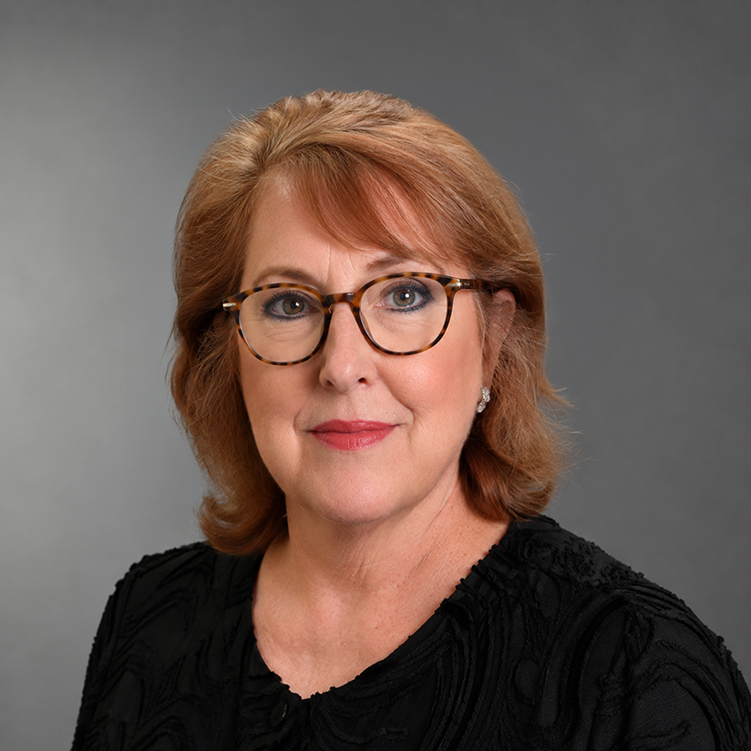 Professional headshot of Judy Wilson wearing tortoiseshell glasses and a black textured top against a gray background.