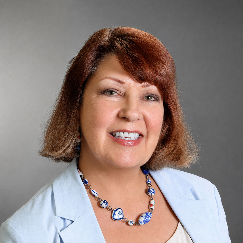 Professional headshot of Monnie Whitson smiling, wearing a light blue blazer and a decorative blue-and-white necklace against a gray background.