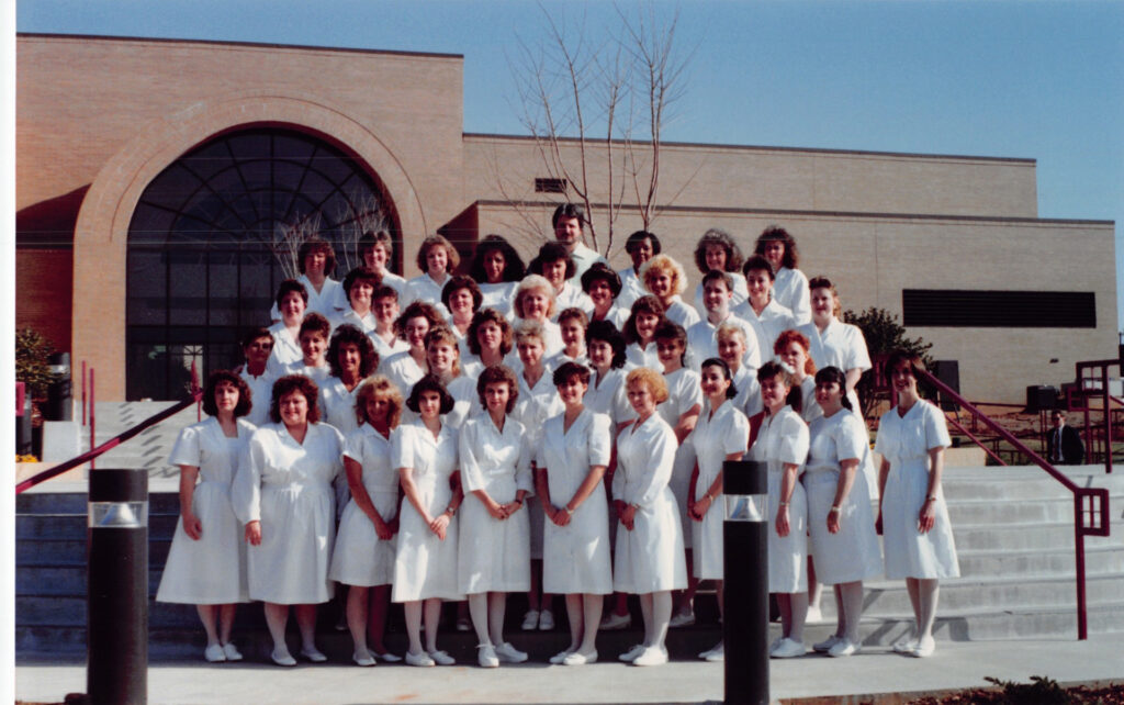 Jeffrey West with fellow Greenville Technical College nursing students in white uniforms, posing for a class photo outside a campus building in the early 1990s.